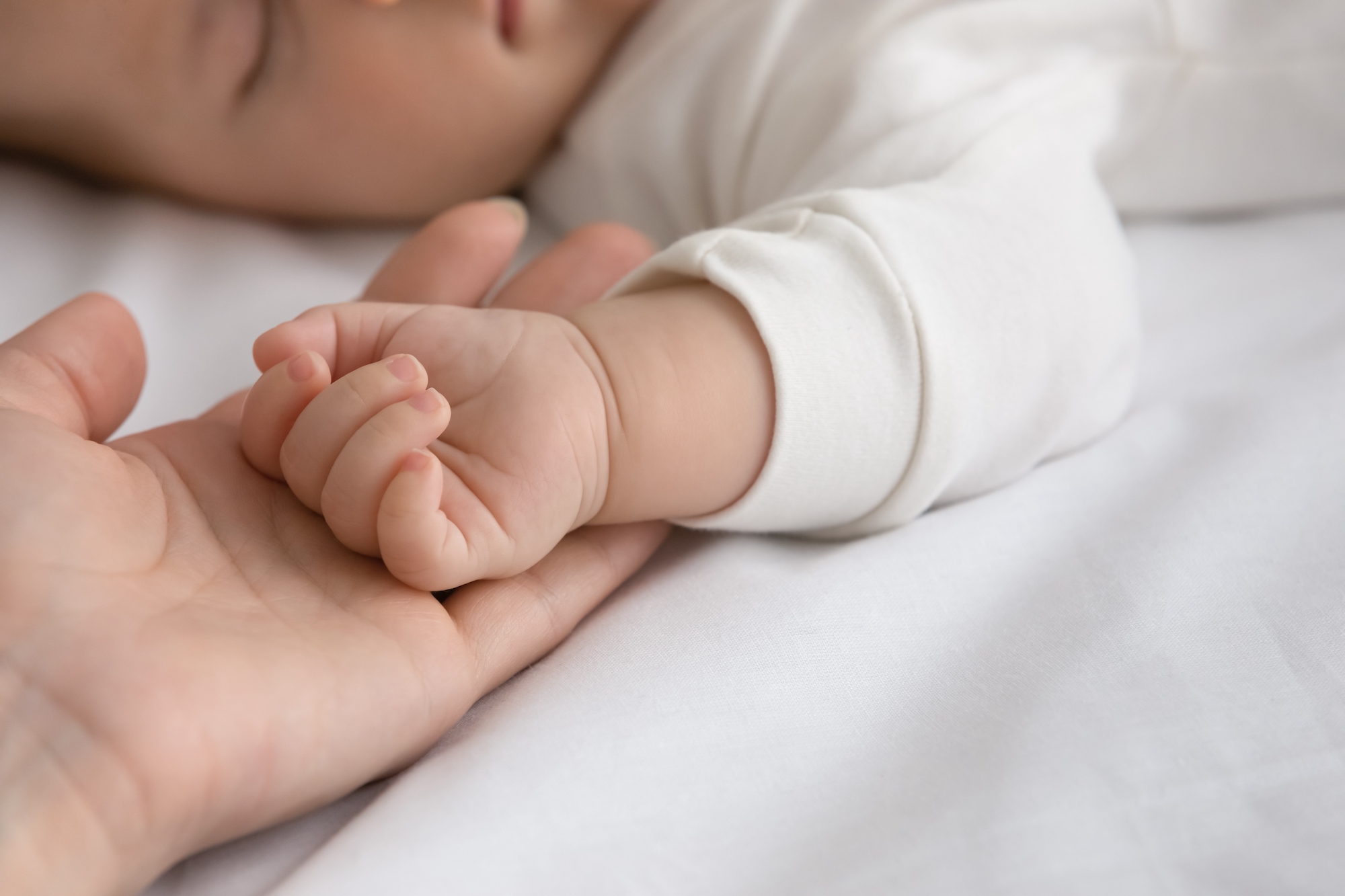 New mom and newborn baby hands close up. Mother holding arm of few month infant son or daughter sleeping on white sheet, touching kid with love and tenderness. Childbirth, motherhood concept. Close up